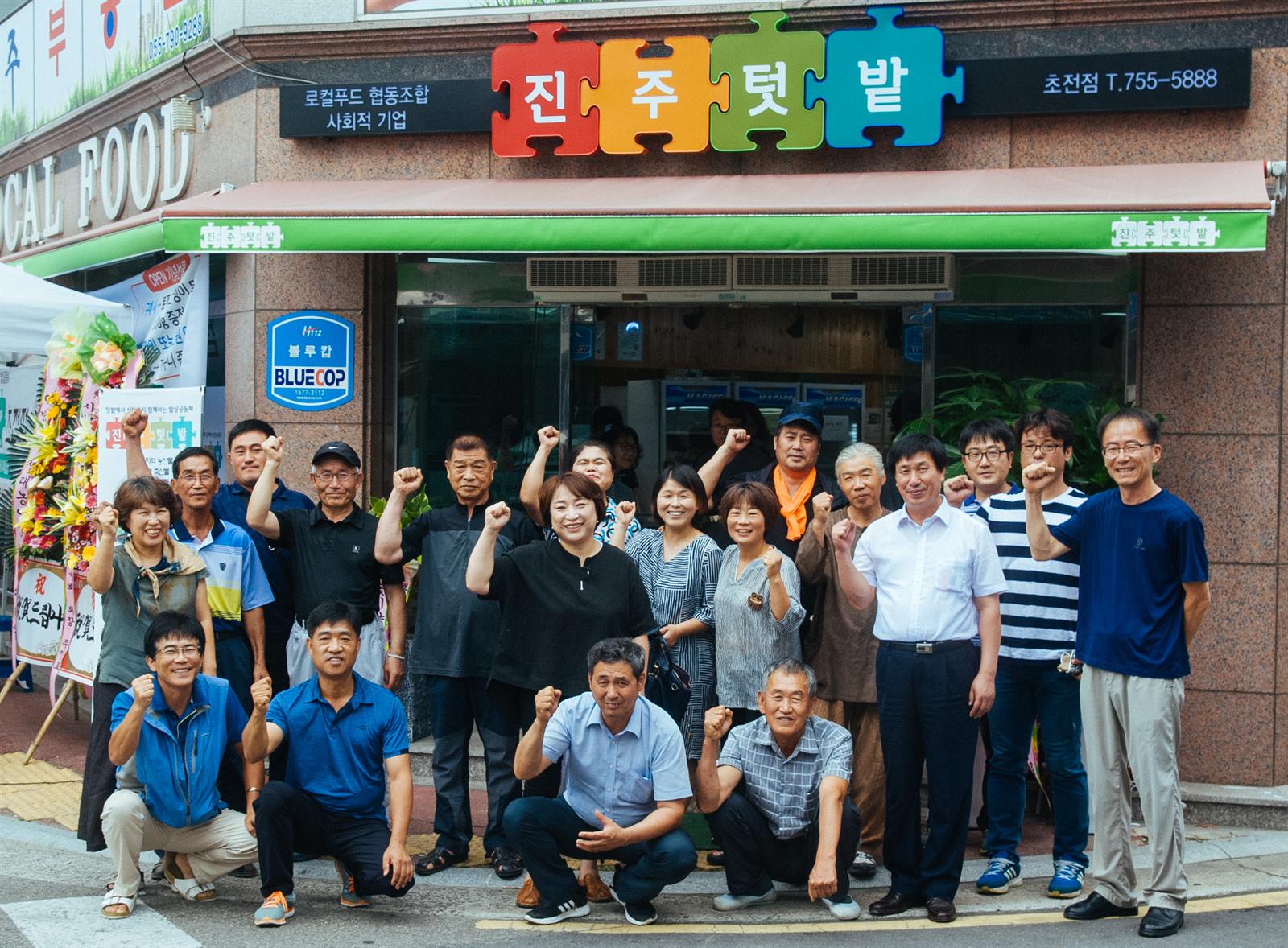 초전점 외관. 진주텃밭 퍼즐 간판이 걸린 매장 앞에서 조합원과 생산자들이 주먹을 들고 함께 사진을 찍고 있다.
